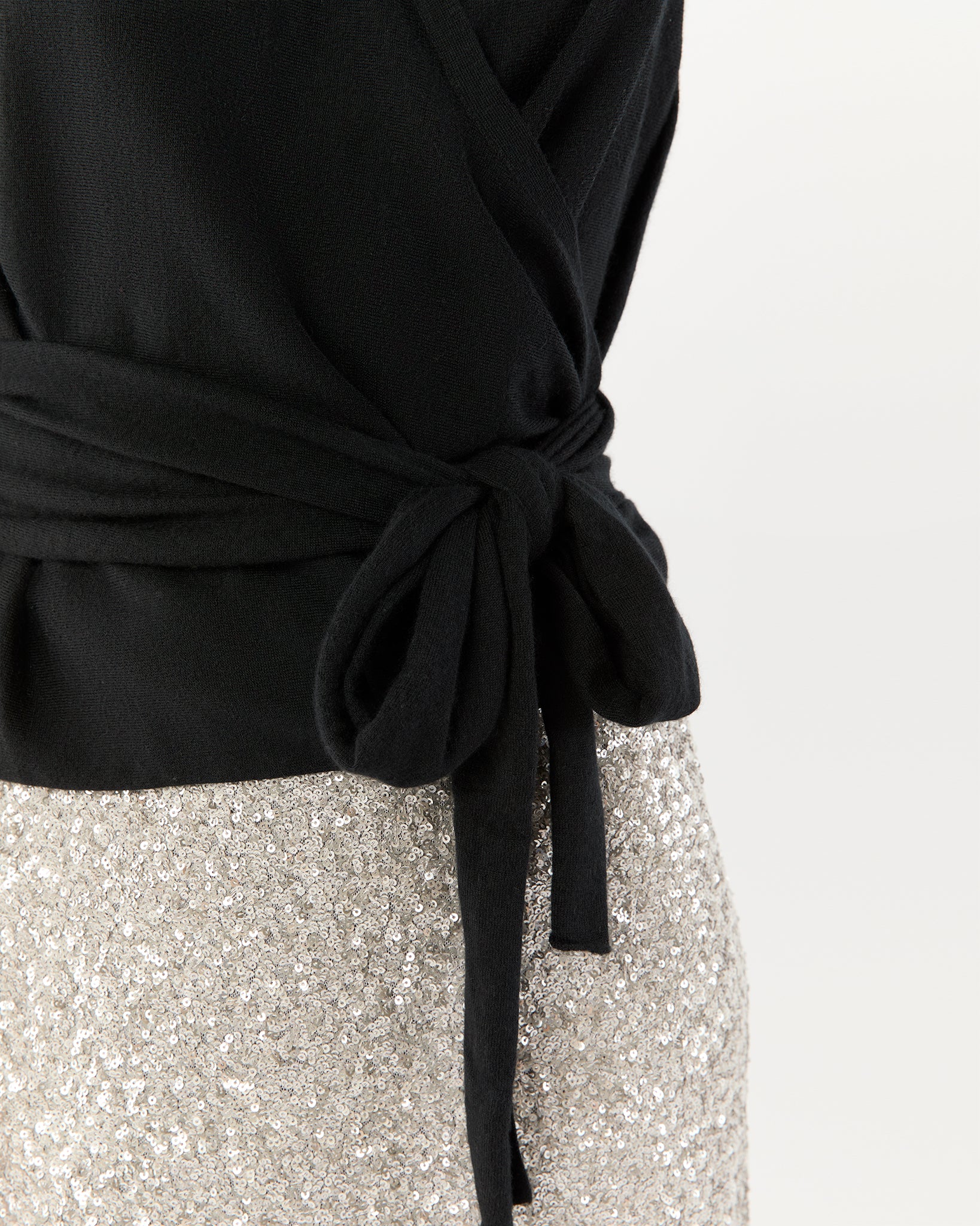 A close-up of a person wearing the Ann Mashburn Mika Cashmere Wrap Sweater in black, tied at the waist, paired with a silver sequin skirt against a plain background.