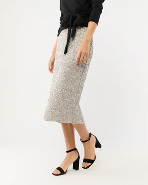 A woman stands against a plain background wearing the Ann Mashburn Sequin Pull-On Skirt with a black long-sleeve top and black ankle-strap block heel sandals.