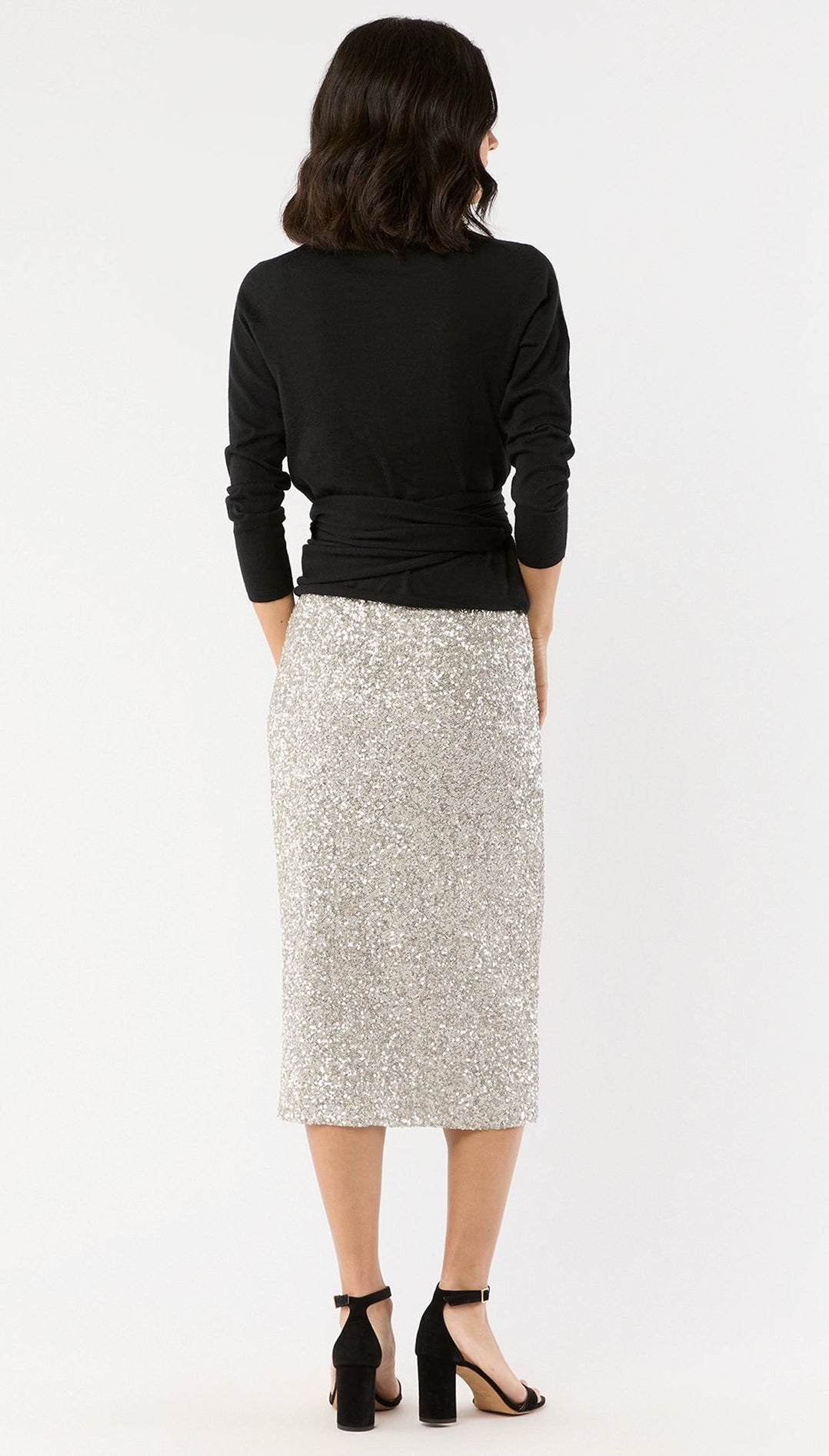 A woman faces away against a white background, wearing an Ann Mashburn Sequin Pull-On Skirt, a black long-sleeve top, and black high-heeled sandals.