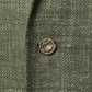 A close-up of the textured green fabric from the Sid Mashburn Ghost Blazer, showcasing a small, uniquely shaped brass button sewn onto it.