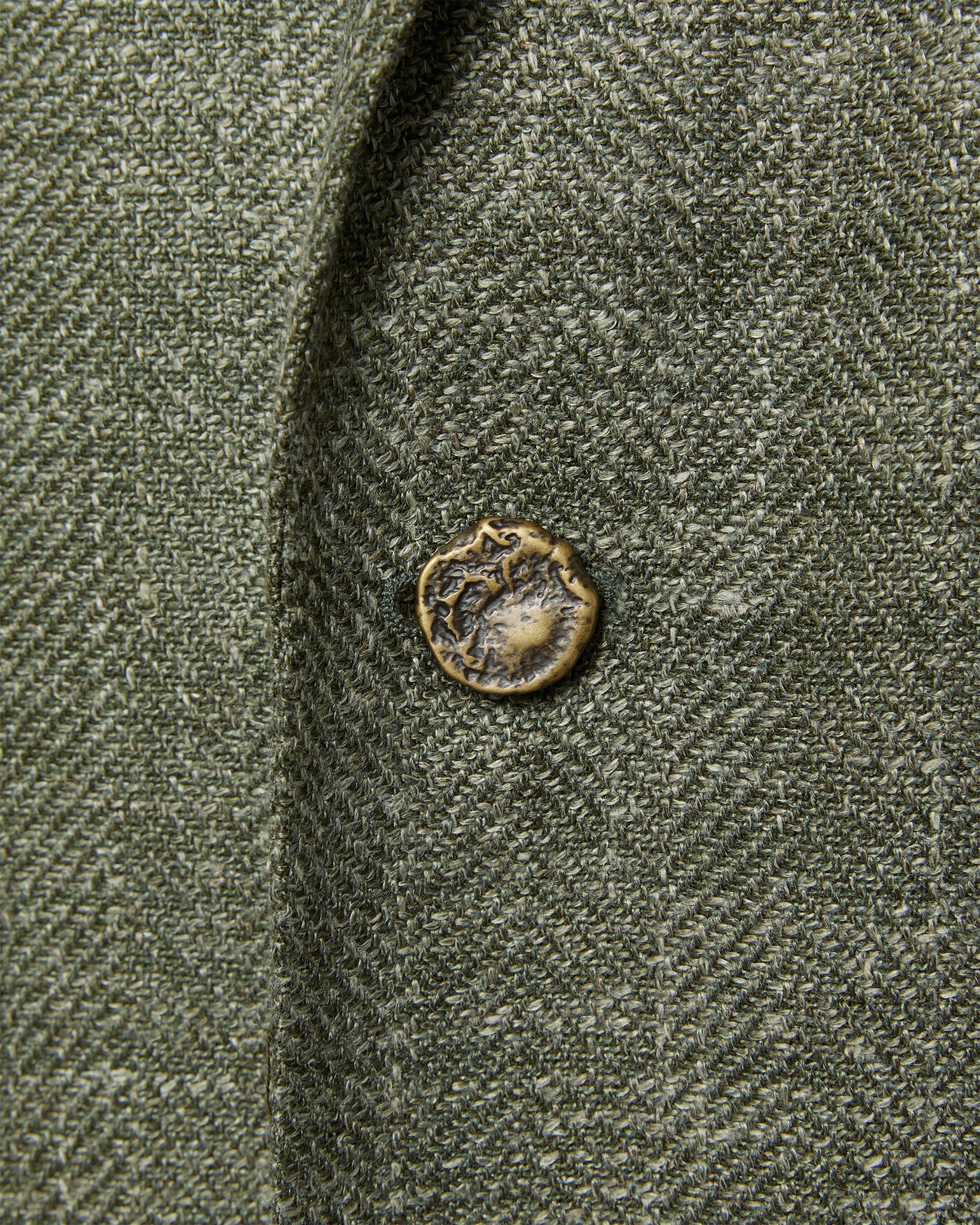 A close-up of the textured green fabric from the Sid Mashburn Ghost Blazer, showcasing a small, uniquely shaped brass button sewn onto it.