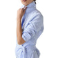 A woman wears the Hilary Wrap Small Bengal Top by Ann Mashburn, a light blue long-sleeve poplin blouse with rolled sleeves and a self-tie belt at the waist, standing against a white background.