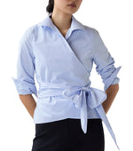 A woman wearing the Ann Mashburn Hilary Wrap Small Bengal Top in light blue poplin with a self-tie waist, paired with black pants.