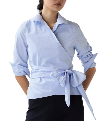 A woman wearing the Ann Mashburn Hilary Wrap Small Bengal Top in light blue poplin with a self-tie waist, paired with black pants.