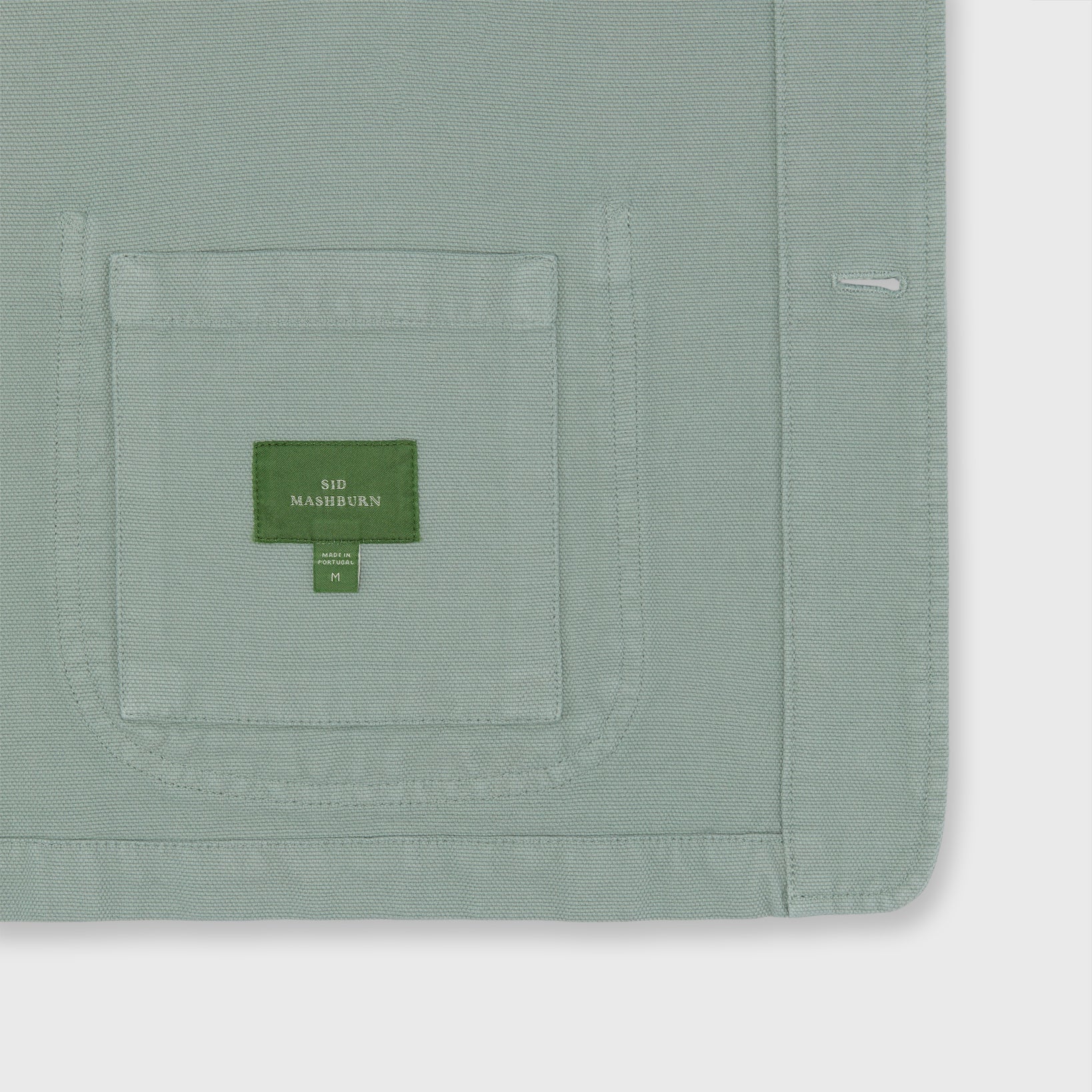 A close-up of a sage green cotton canvas fabric showcases a stitched pocket with a green Sid Mashburn label and white sizing text—signature details of the classic Sid Mashburn Chore Jacket.