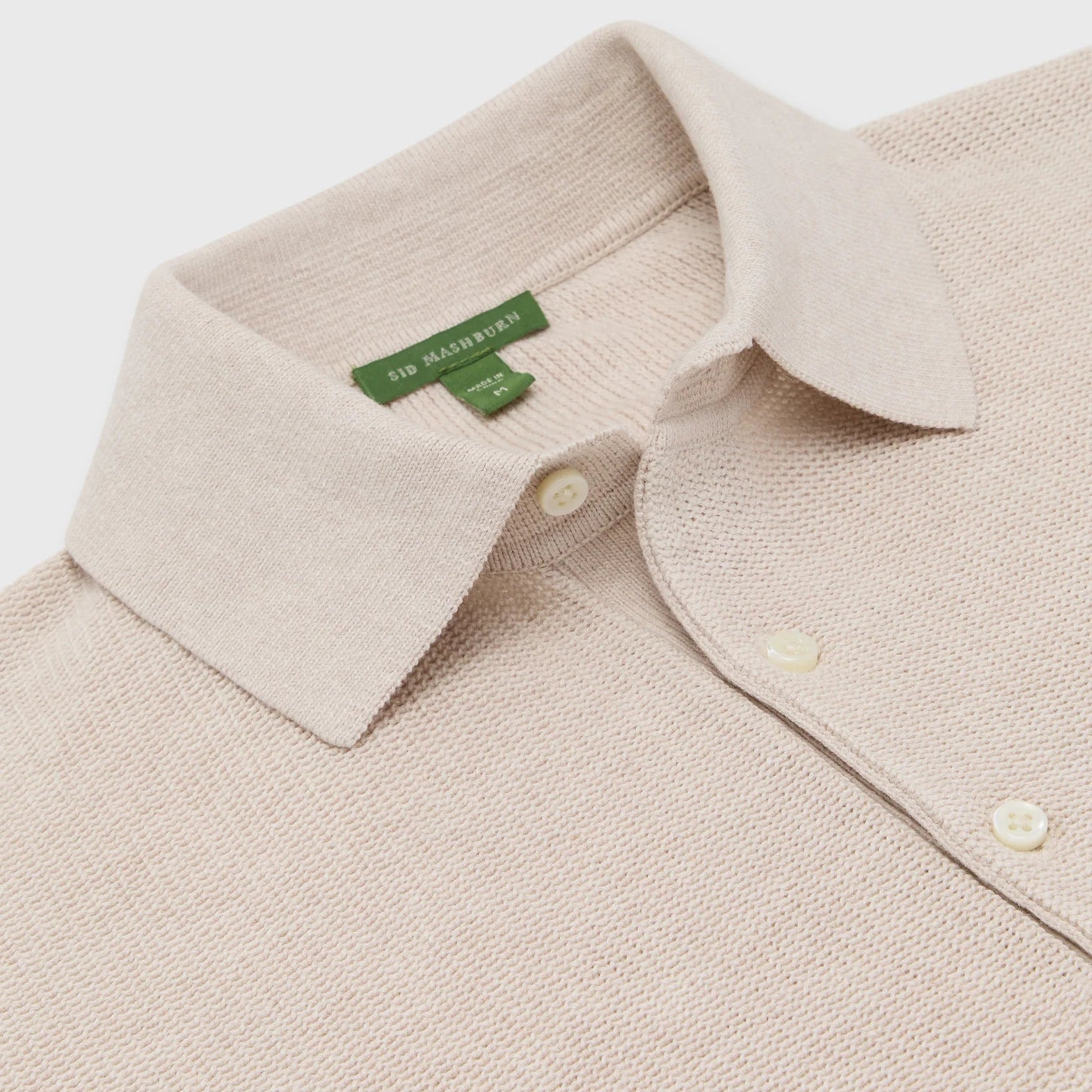The Sid Mashburn Textured Hyannis Polo Sweater in beige cotton-linen features a pointed collar, cream buttons on a French placket, and a green brand label inside the collar, shown flat to highlight the upper chest and collar.