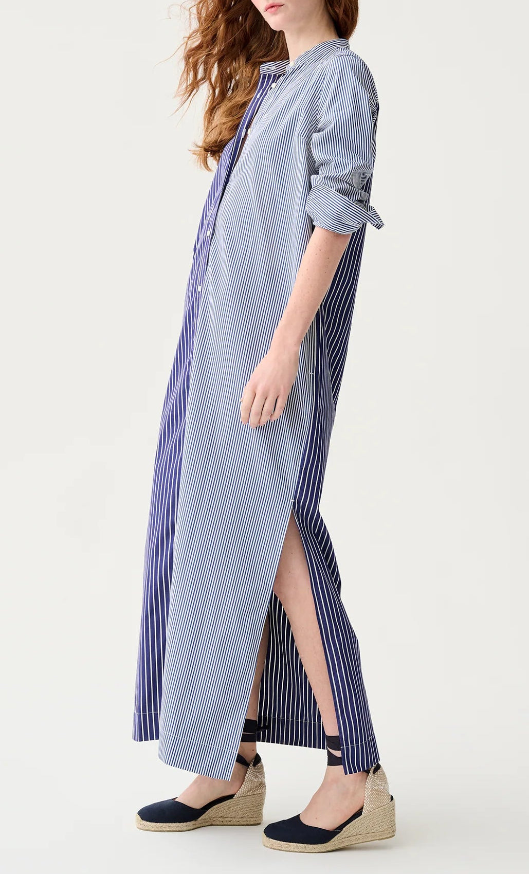 A woman in the Ann Mashburn Mandarin Talitha Mixed Stripe Poplin Shirtdress—a long blue and white striped shirtdress with rolled sleeves—wears espadrille wedges and stands against a plain background.