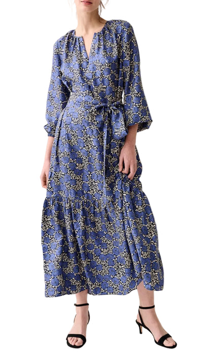 A woman wearing the Aba Printed Silk Twill Dress by Ann Mashburn—a long-sleeved blue floral wrap dress with elasticated cuffs, a self-tie waist belt—paired with black strappy heels.