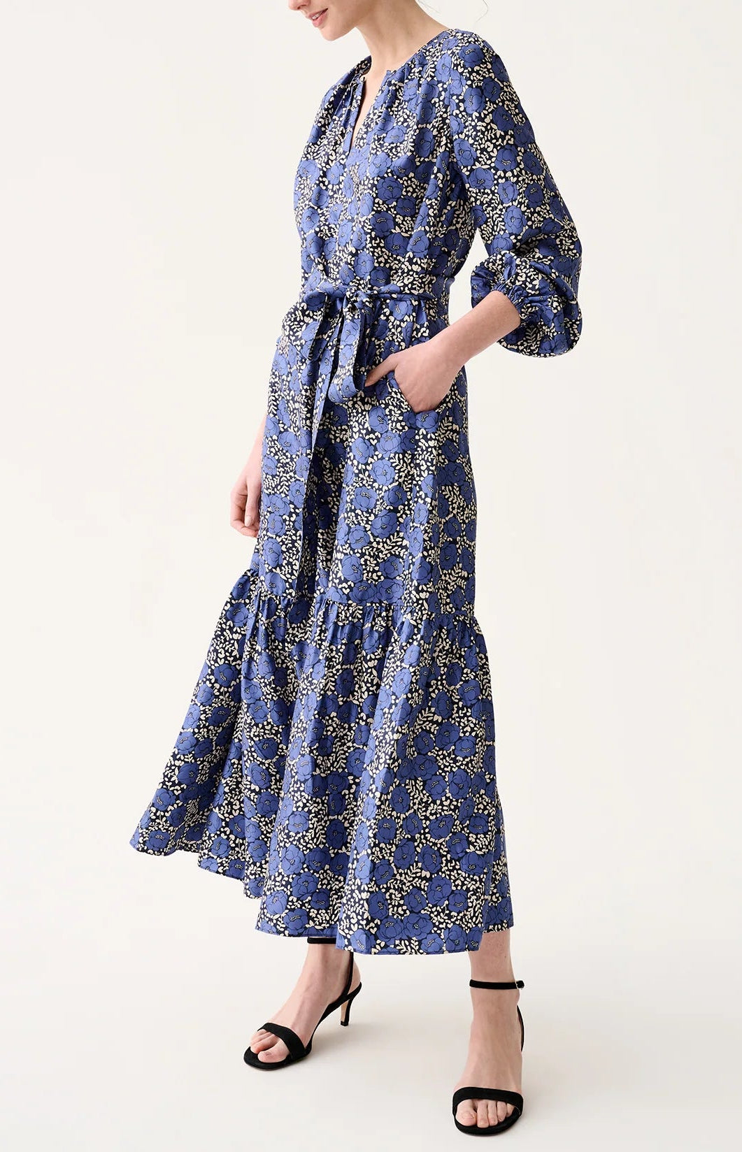 A person wearing the Aba Printed Silk Twill Dress by Ann Mashburn—featuring long sleeves, a blue floral print, elasticated cuffs, waist tie, and tiered skirt—paired with black strappy heels.