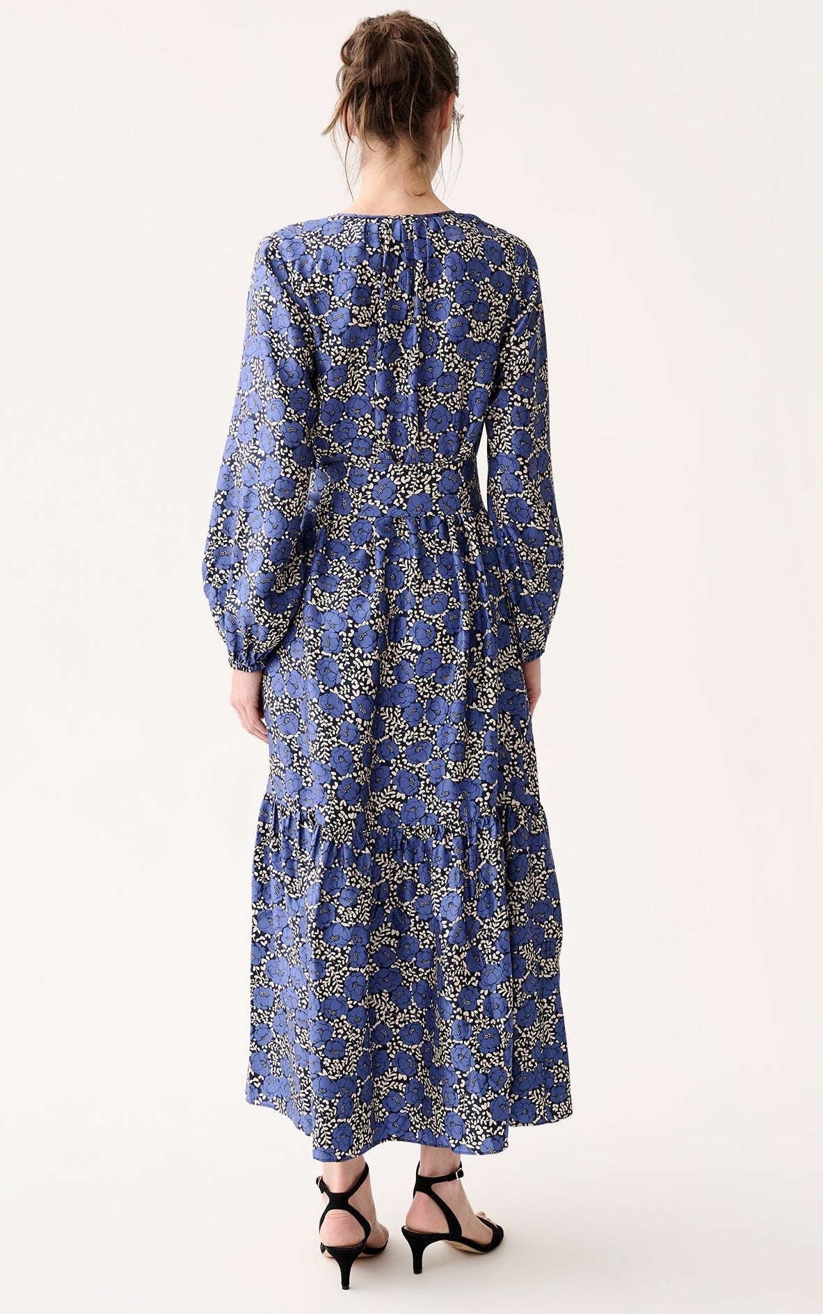 A woman faces away, wearing the Ann Mashburn Aba Printed Silk Twill Dress—an ankle-length, long-sleeved blue floral dress with elasticated cuffs and a self-tie belt—paired with black strappy sandals against a white background.