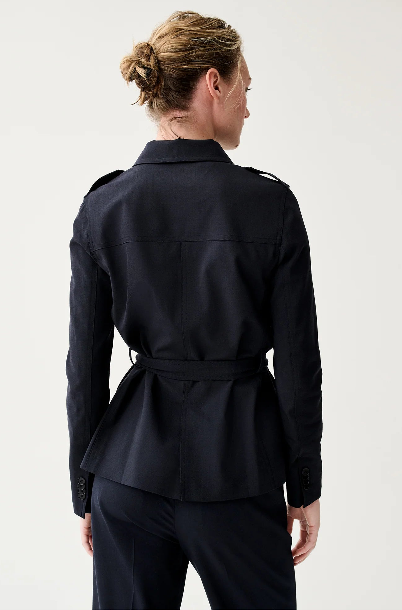 A person with light hair in a bun is seen from behind, wearing Ann Mashburn's Safari Bi-Stretch Tropical Wool Jacket with matching trousers against a plain background.