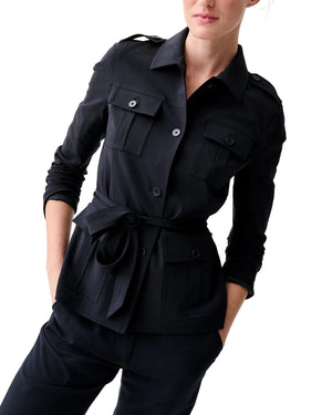 A woman stands confidently with her hands in her pockets, styled in the Ann Mashburn Safari Bi-Stretch Tropical Wool Jacket—belted, long-sleeve, black with buttoned pockets—paired with matching black pants as a chic third piece.