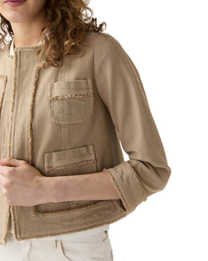A woman wears the Ann Mashburn Kiki Stretch Canvas Jacket in beige with frayed edges and front pockets, paired with white pants, set against a white background.