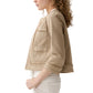 A woman with curly hair is shown in profile wearing the Ann Mashburn Kiki Stretch Canvas Jacket—a light beige cropped jacket with front pockets—paired with light-colored pants, set against a white background.