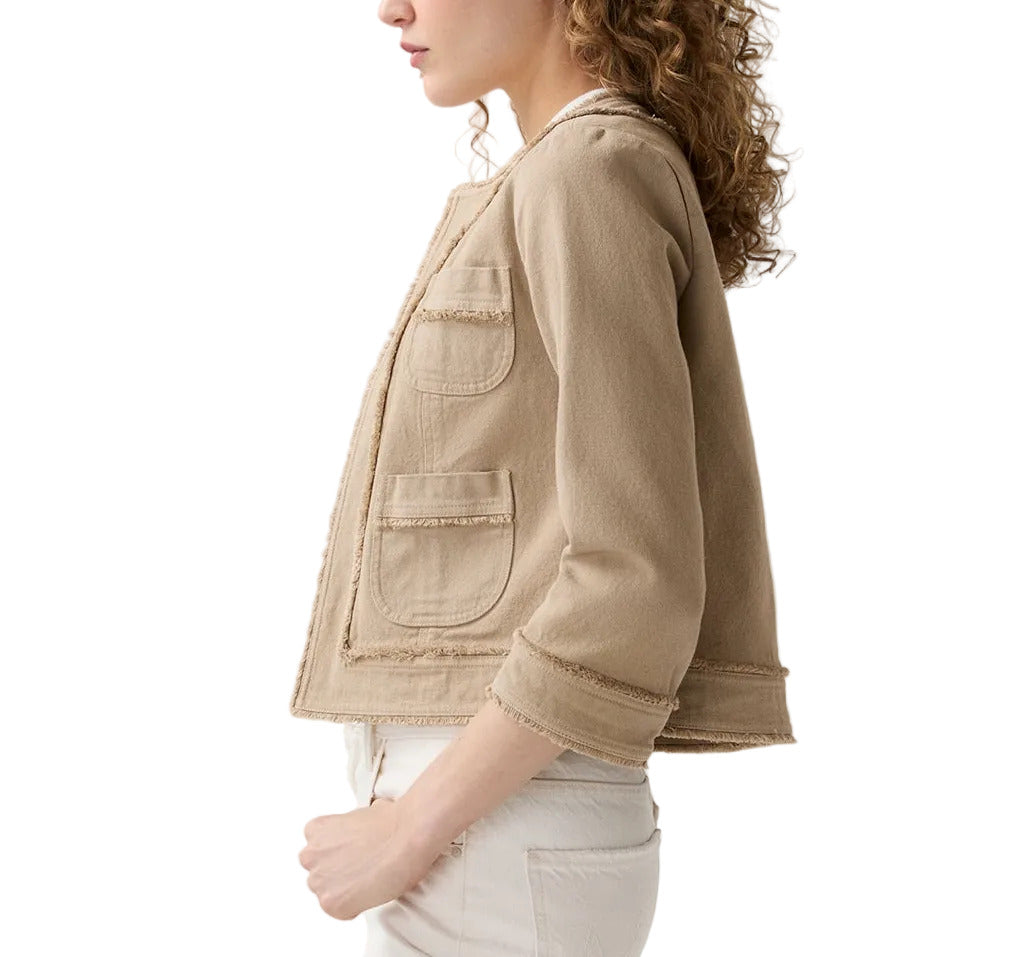 A woman with curly hair is shown in profile wearing the Ann Mashburn Kiki Stretch Canvas Jacket—a light beige cropped jacket with front pockets—paired with light-colored pants, set against a white background.