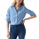 A woman stands against a plain background wearing the Ann Mashburn Sidney Stripe Shirt—a blue and white button-up with rolled sleeves, spread collar—and blue jeans.