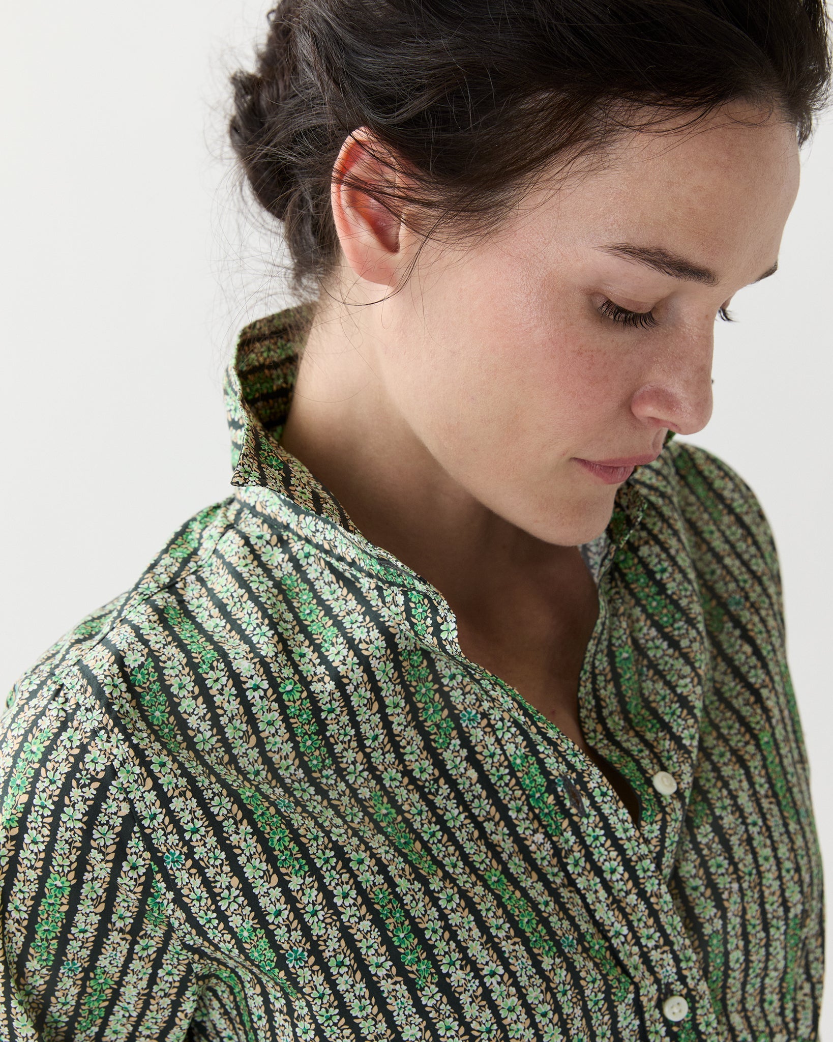 A woman with dark hair in a loose bun looks down, wearing the Ann Mashburn Classic Shirtwaist Floral Stripe Dress with white buttons against a plain background.