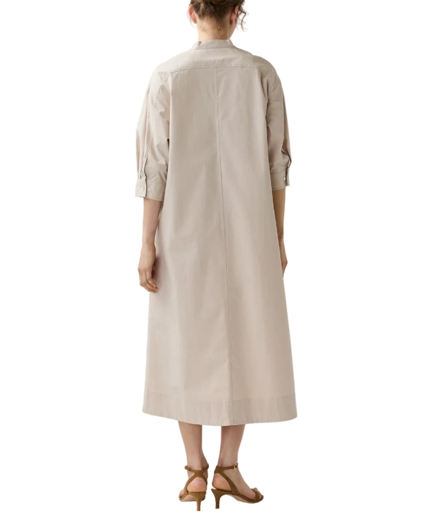 A person stands facing away, wearing the Cadie Paperweight Stretch Poplin Dress by Ann Mashburn in light beige with rolled-up sleeves and tan heeled sandals.