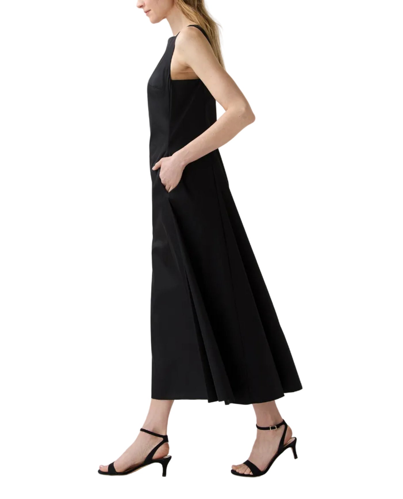 A woman stands in profile with her hands in the pockets of the Ann Mashburn Rosalind Dress, a sleeveless black poplin style, paired with black heeled sandals.