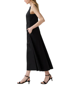 A woman stands in profile with her hands in the pockets of the Ann Mashburn Rosalind Dress, a sleeveless black poplin style, paired with black heeled sandals.