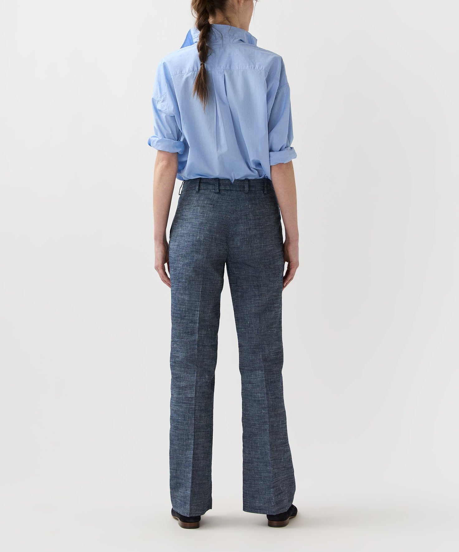 Person standing facing away, wearing high-waisted Lane Cotolino Chambray Pant by Ann Mashburn and a light blue button-up shirt with rolled sleeves against a plain background.
