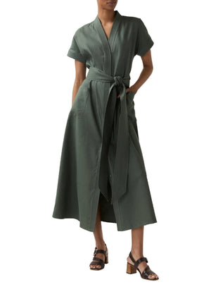 A person wearing the Ann Mashburn Lianna Stretch Cotolino Dress, an olive green short-sleeved shirtdress with a tie belt, styled with black heeled sandals and standing with hands in pockets.