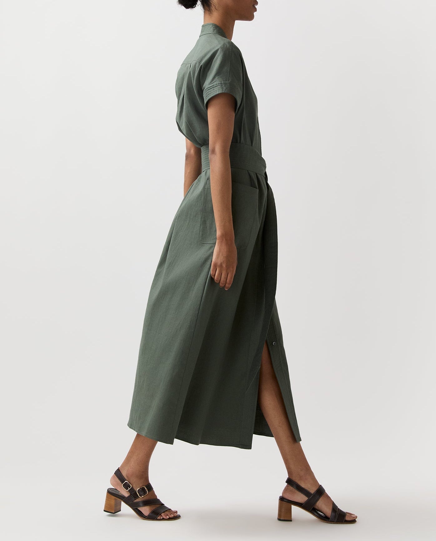 A person in profile wears the Ann Mashburn Lianna Stretch Cotolino Dress—belted, short-sleeve, olive green with an A-line skirt—paired with black heeled sandals, set against a plain background.