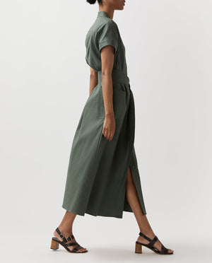 A person in profile wears the Ann Mashburn Lianna Stretch Cotolino Dress—belted, short-sleeve, olive green with an A-line skirt—paired with black heeled sandals, set against a plain background.