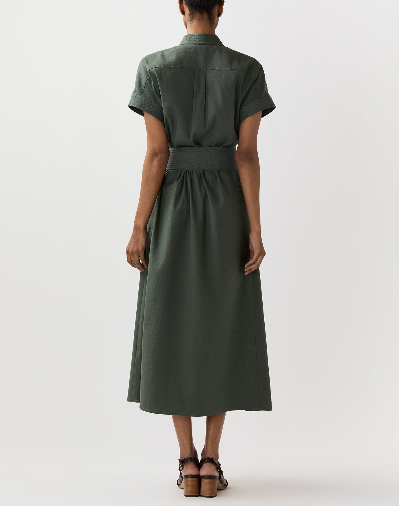 A person faces away, wearing the Ann Mashburn Lianna Stretch Cotolino Dress—short-sleeved, belted, with an A-line skirt in forest green—and brown heeled sandals against a plain white background.