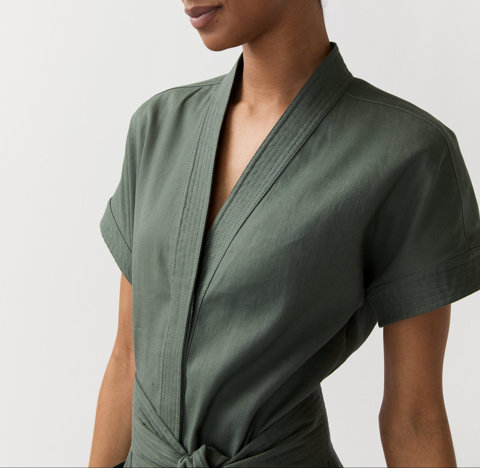 A person wearing the Ann Mashburn Lianna Stretch Cotolino Dress in olive green, featuring a deep V-neck, tied waist, and A-line skirt, photographed from the chest up against a plain background.