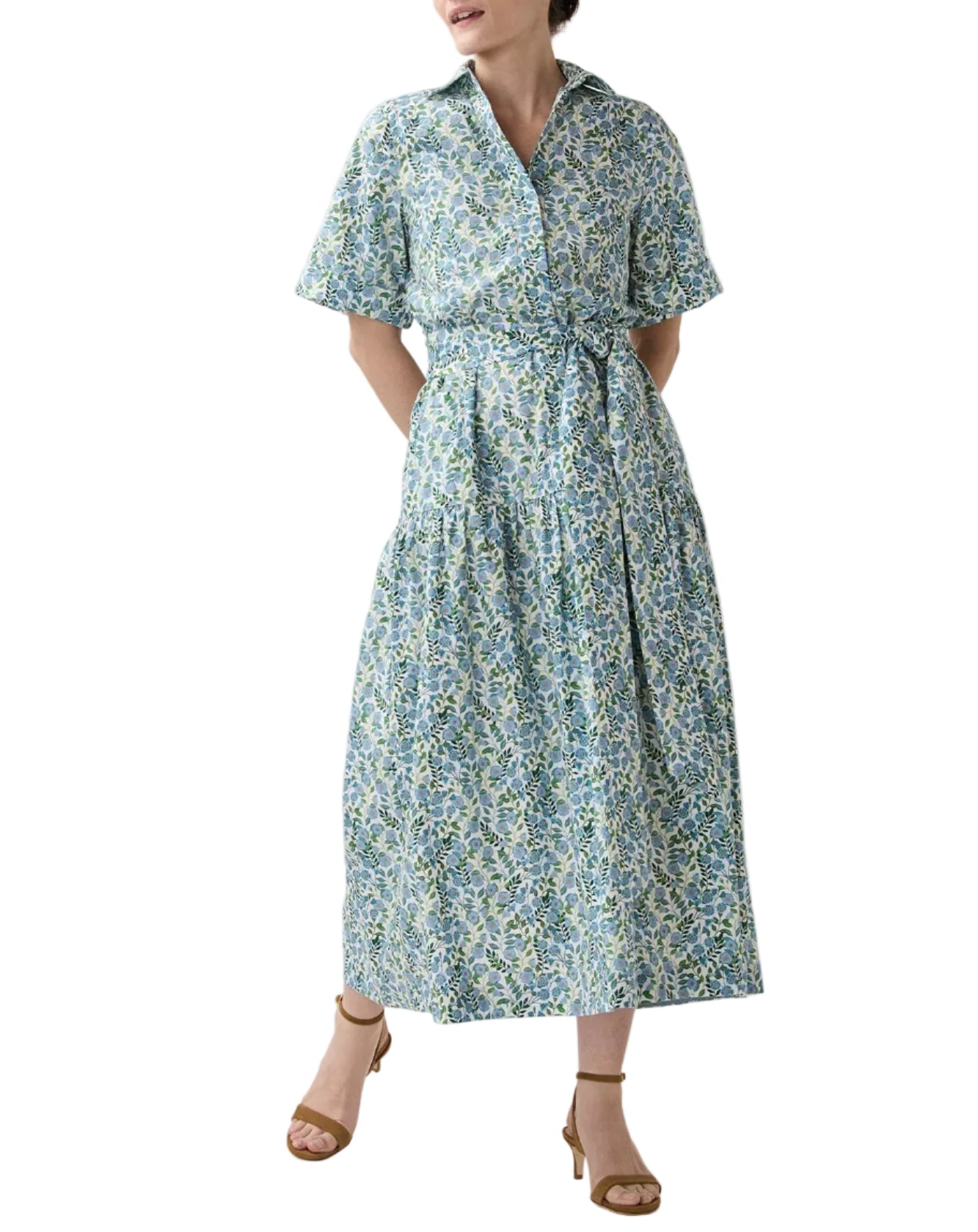 A person wears Ann Mashburn's Nathalie Vigers' Vine Liberty Dress, a short-sleeve, collared cotton shirtdress in blue and green florals with a tied waist and tiered skirt, paired with tan heeled sandals.