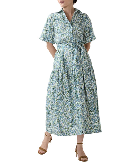 A person wears Ann Mashburn's Nathalie Vigers' Vine Liberty Dress, a short-sleeve, collared cotton shirtdress in blue and green florals with a tied waist and tiered skirt, paired with tan heeled sandals.