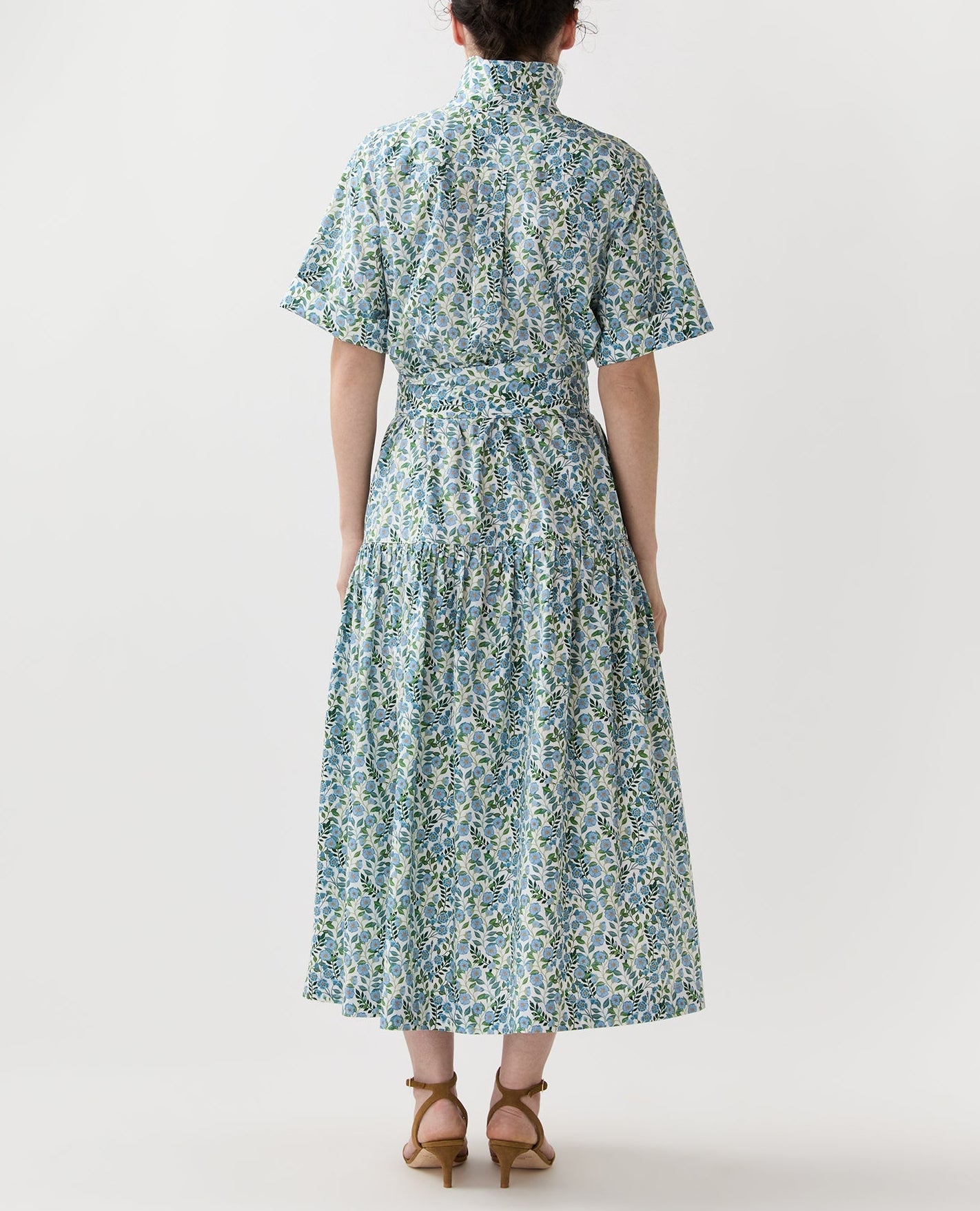 A person stands facing away, wearing the Nathalie Vigers' Vine Liberty Dress by Ann Mashburn—blue and green floral cotton with short sleeves and a belted waist—paired with tan heeled sandals.