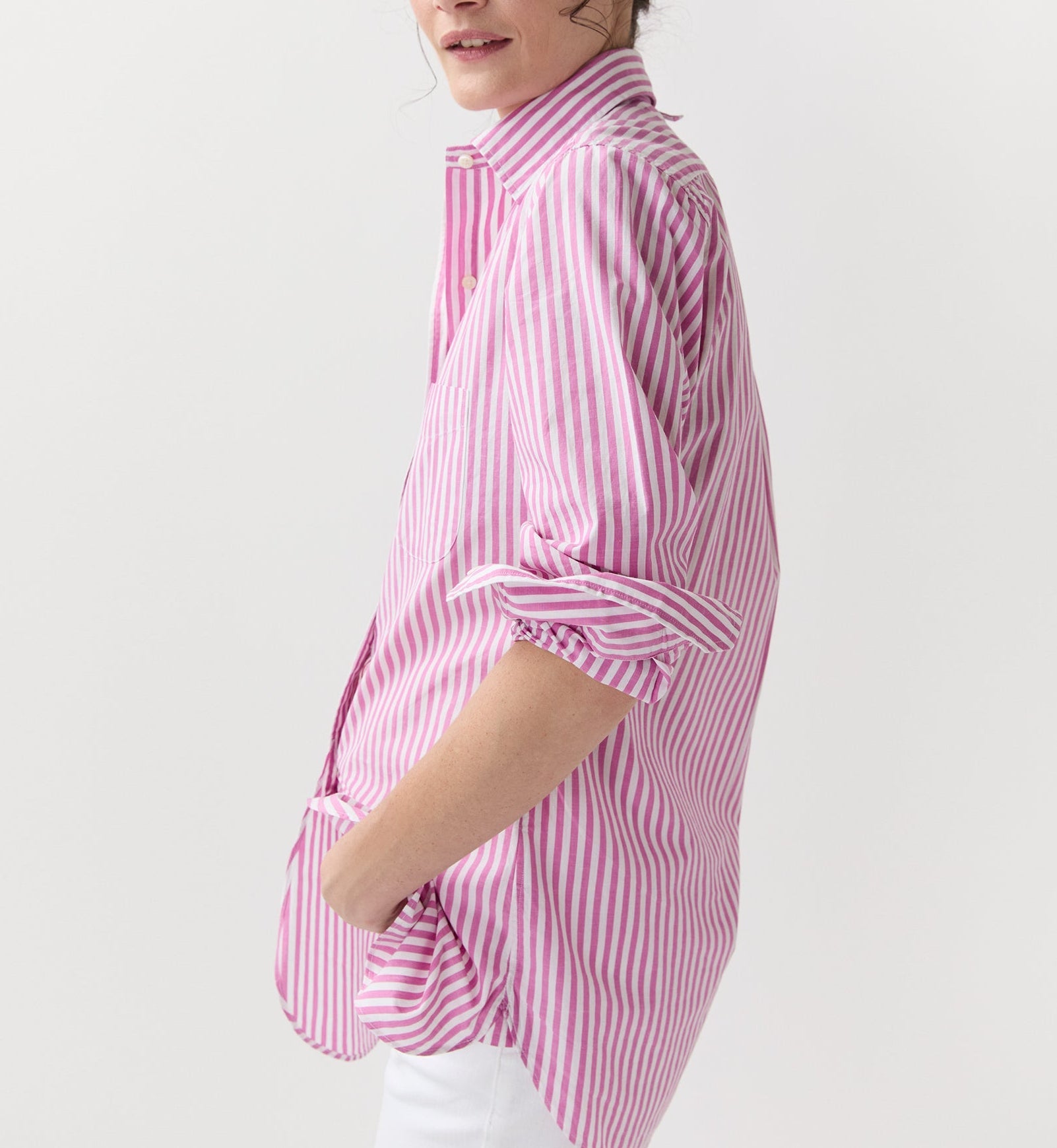 A person in the Ann Mashburn Boyfriend Stripe Chambray Shirt, a pink and white striped long-sleeve women's top, styled with white pants and standing sideways with one hand in their pocket.