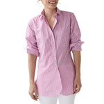 A woman smiles, facing slightly to the side, in the Ann Mashburn Boyfriend Stripe Chambray Shirt—pink and white stripes in soft Italian chambray with rolled-up sleeves—paired with white pants.