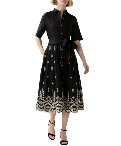 A woman wears the Ann Mashburn Short-Sleeve Classic Shirtwaist Dress, featuring black cotton chambray, a tied waist, and white floral embroidery on the skirt and hem.