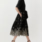 A woman in the Ann Mashburn Short-Sleeve Classic Shirtwaist Dress, featuring black cotton chambray and white floral embroidery at the hem, pairs it with black heeled sandals while walking against a plain background.