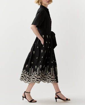 A woman in the Ann Mashburn Short-Sleeve Classic Shirtwaist Dress, featuring black cotton chambray and white floral embroidery at the hem, pairs it with black heeled sandals while walking against a plain background.