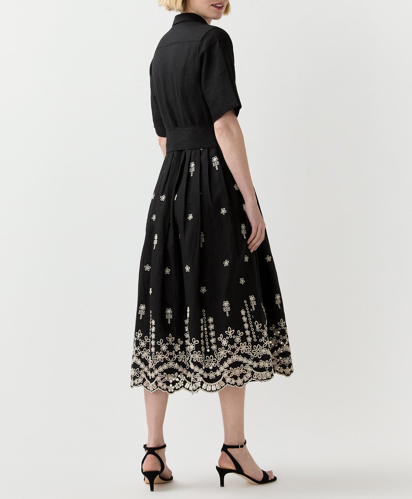 A woman stands facing away, wearing the Ann Mashburn Short-Sleeve Classic Shirtwaist Dress with white floral embroidery and black heeled sandals.