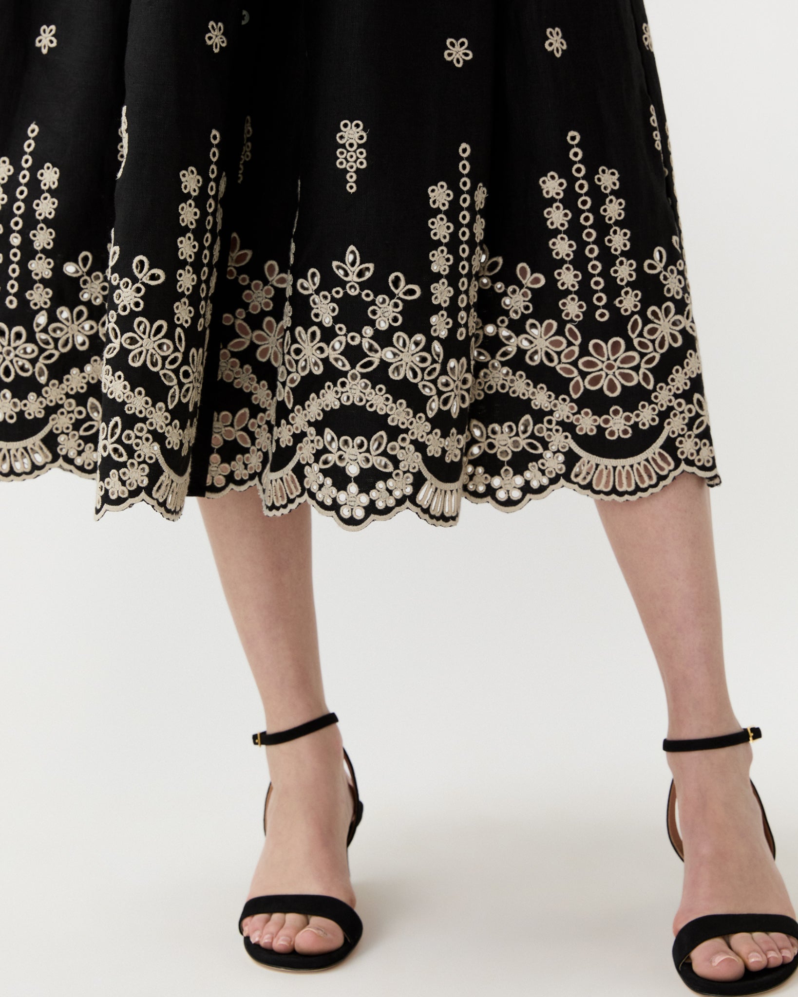 A person in Ann Mashburn's Short-Sleeve Classic Shirtwaist Dress—a black skirt with white floral embroidery near the hem—styled with black ankle-strap sandals for a timeless, feminine look.