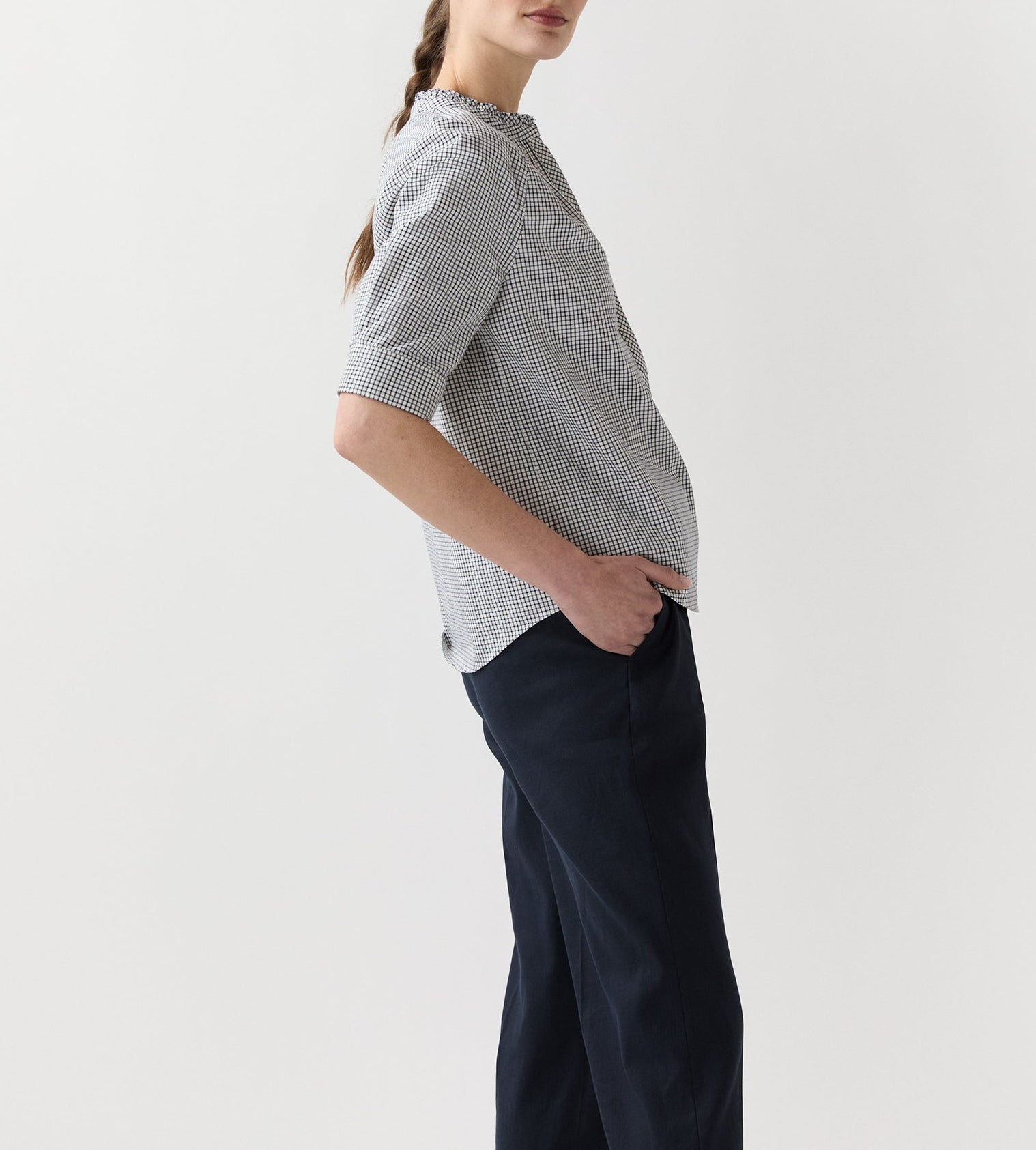 A person stands sideways with one hand in their pocket, wearing the Sage Blouse by Ann Mashburn and dark pants against a plain background.