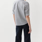 A person stands facing away, wearing the Ann Mashburn Sage Blouse—a checked poplin top with elbow sleeves—and dark trousers against a plain white background.