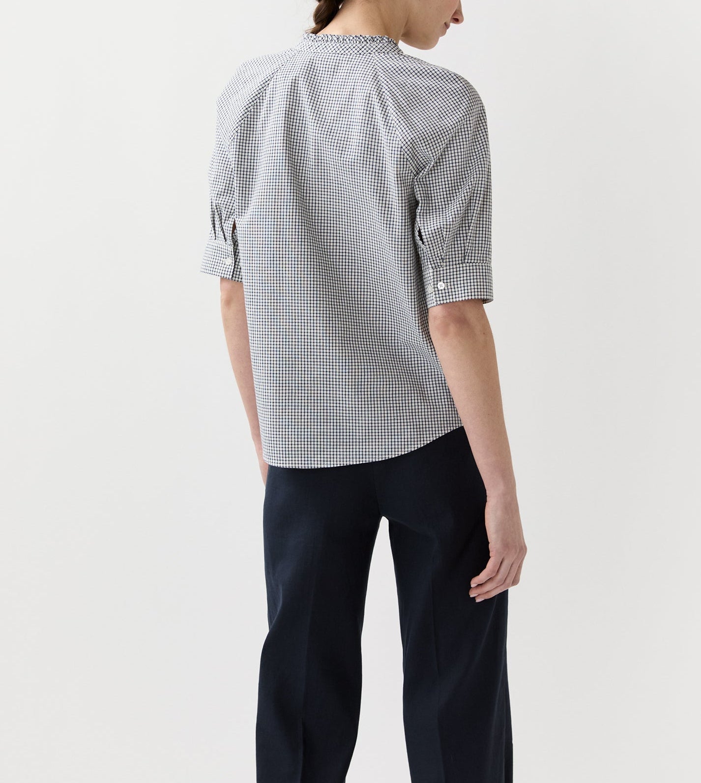 A person stands facing away, wearing the Ann Mashburn Sage Blouse—a checked poplin top with elbow sleeves—and dark trousers against a plain white background.