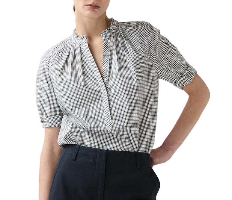 A person wearing the Ann Mashburn Sage Blouse—a black and white checkered Portuguese poplin popover with short sleeves—and dark trousers, standing with one hand in their pocket.