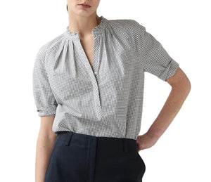 A person wearing the Ann Mashburn Sage Blouse—a black and white checkered Portuguese poplin popover with short sleeves—and dark trousers, standing with one hand in their pocket.