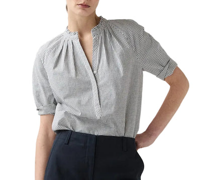 A person wearing the Ann Mashburn Sage Blouse—a black and white checkered Portuguese poplin popover with short sleeves—and dark trousers, standing with one hand in their pocket.