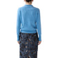 A woman stands facing away against a white background, wearing the Ann Mashburn Connor Cotton/Silk Sweater in light blue paired with a dark blue floral-patterned skirt.