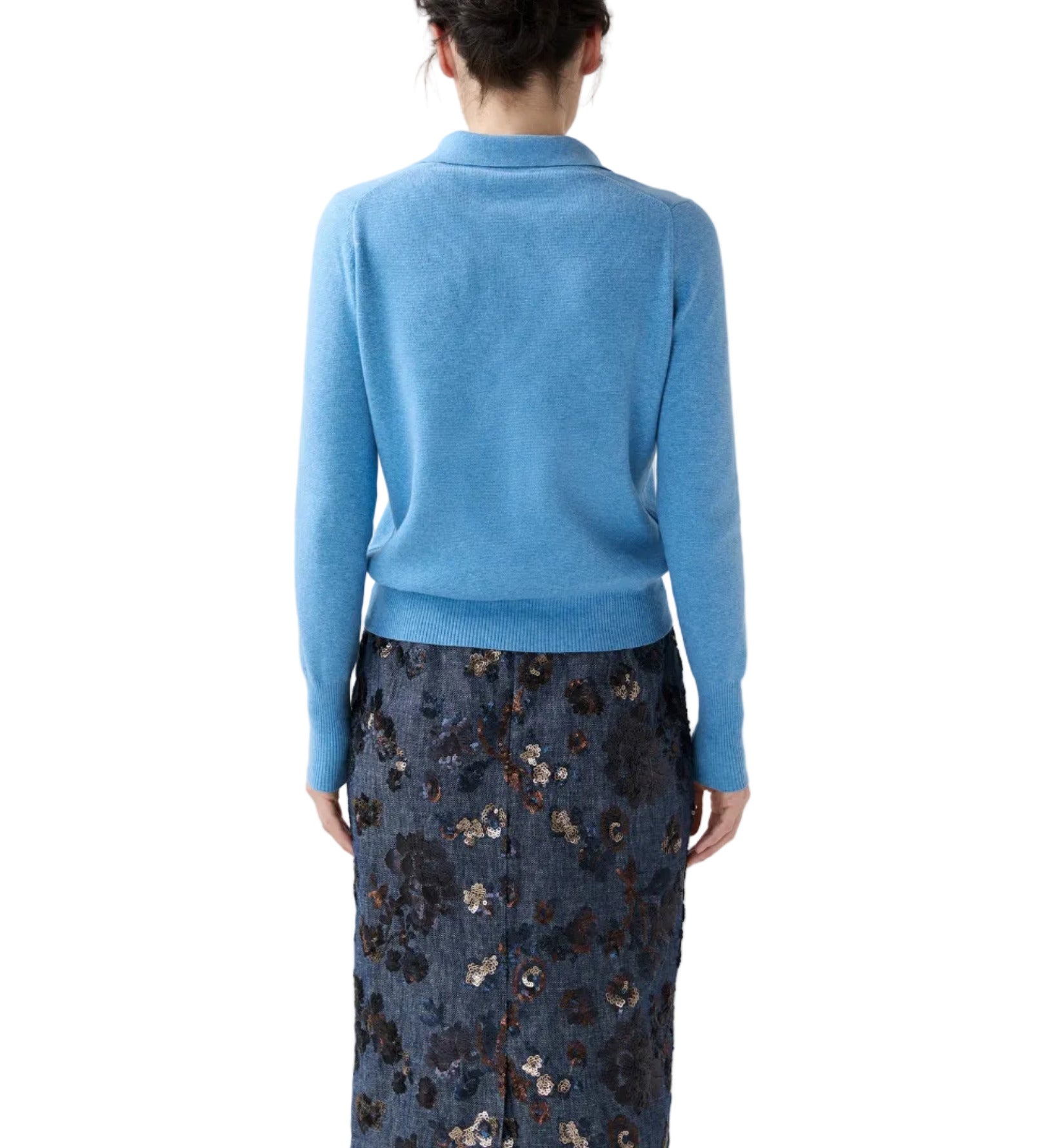 A woman stands facing away against a white background, wearing the Ann Mashburn Connor Cotton/Silk Sweater in light blue paired with a dark blue floral-patterned skirt.
