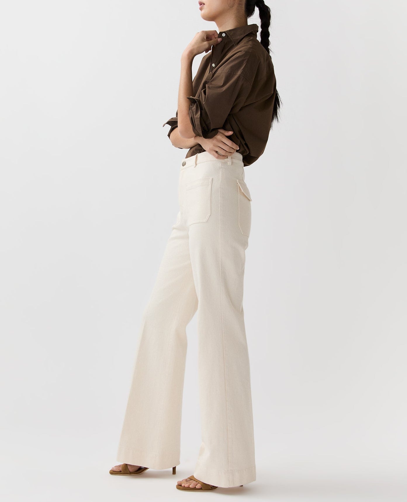 A woman in profile wears a brown long-sleeve shirt, Ann Mashburn Camille Patch-Pocket Stretch Jeans, and heeled sandals against a plain light background.
