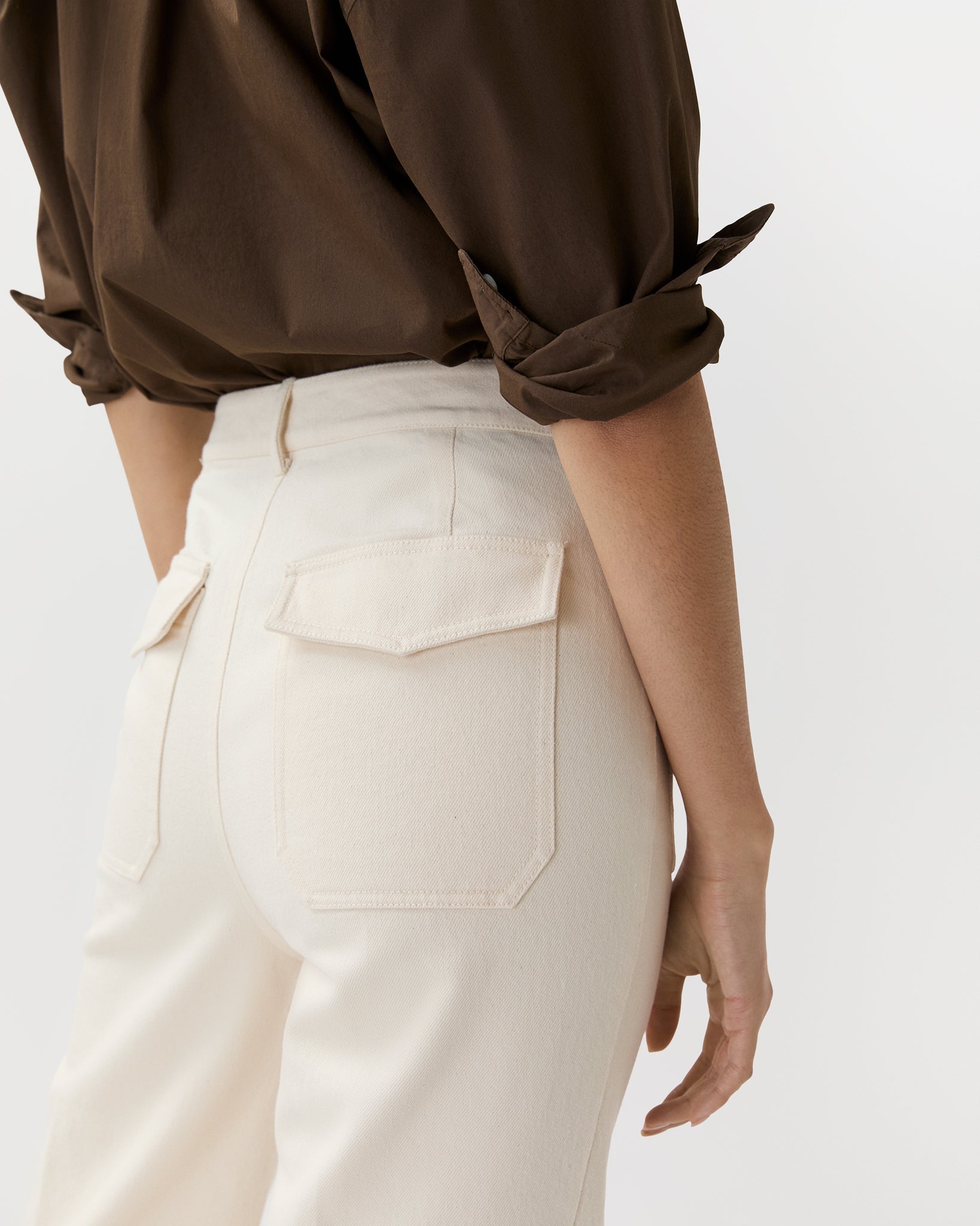 Wearing Ann Mashburn’s Camille Patch-Pocket Stretch Jean, a high-waisted cream-colored style with back flap pockets, paired with a brown rolled-sleeve shirt against a plain background.
